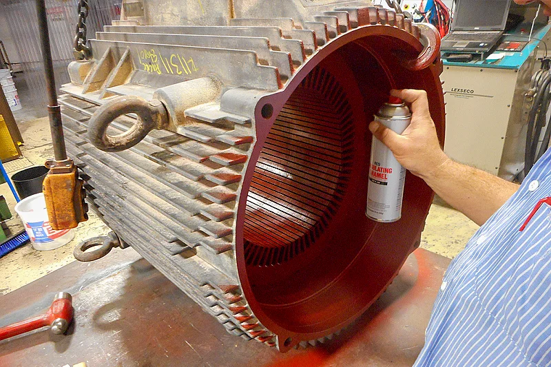 Technician applying red insulating enamel coating to motor stator core to restore dielectric strength before rewind.