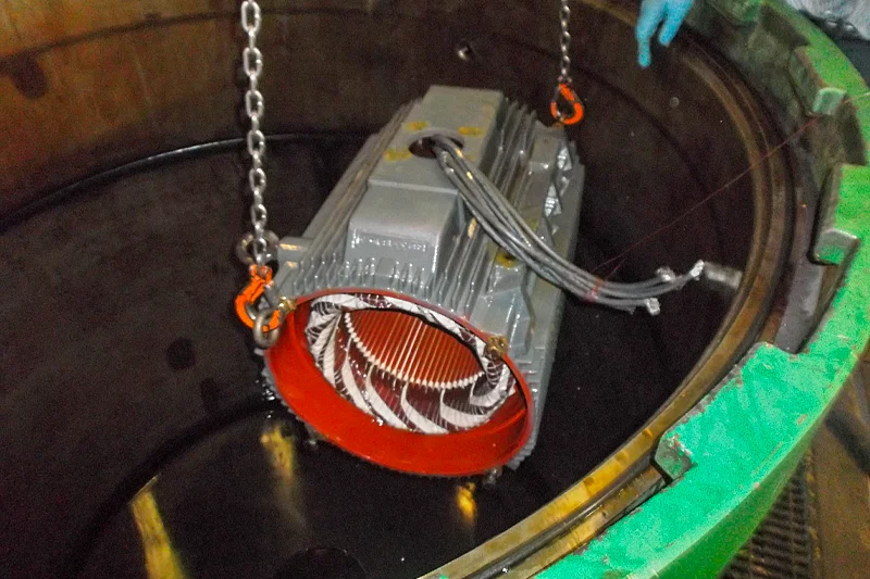 An electric motor stator being lowered into a large vacuum pressure impregnation (VPI) tank filled with resin. Chains and hooks are attached for safe handling.