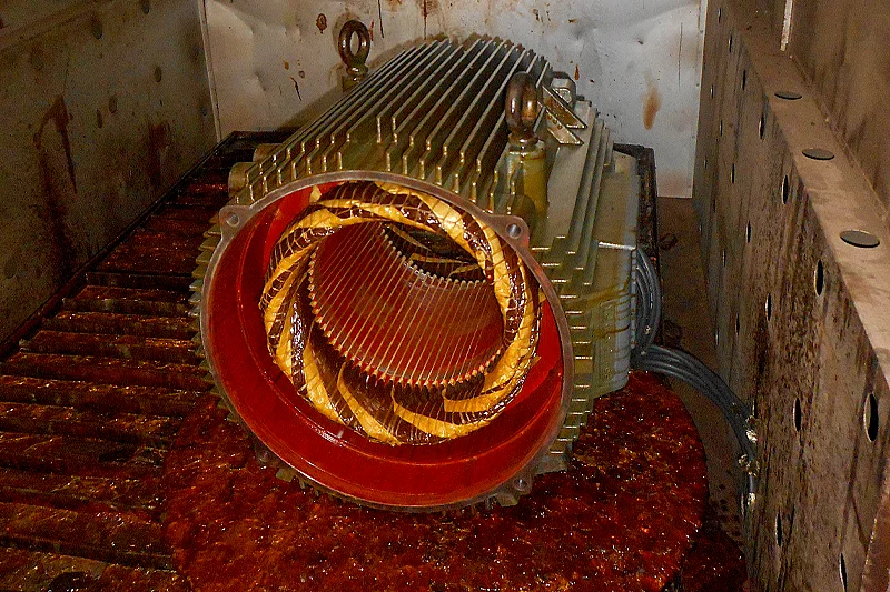A stator sits inside an industrial baking oven, coated with cured resin that gives the windings a glossy finish. The resin has begun to pool at the base, indicating the curing process is underway.