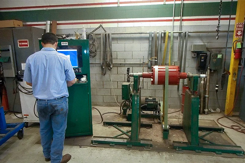 A technician operates a dynamic balancing machine as a red-painted rotor spins between two green support stands. The control cabinet displays vibration data to ensure the rotor meets industry balance standards.